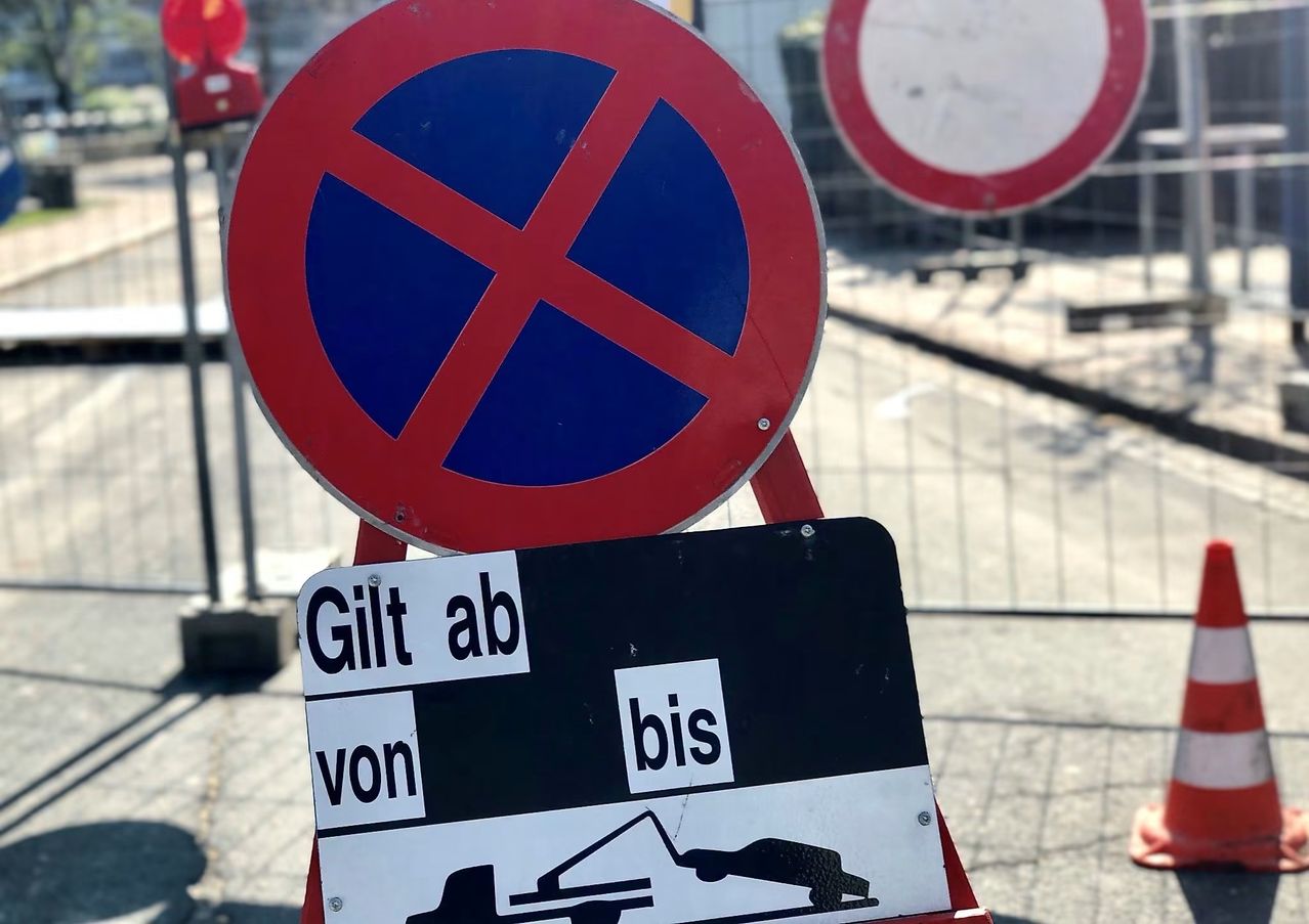 Ein Straßenschild zeigt 'Gültig ab' und 'Gültig bis' mit einem durchgestrichenen Symbol an. Dahinter ist ein großes rotes Schild mit einem blauen X zu sehen, und es gibt eine verschwommene Ansicht eines Metallzauns und eines Gebäudes.