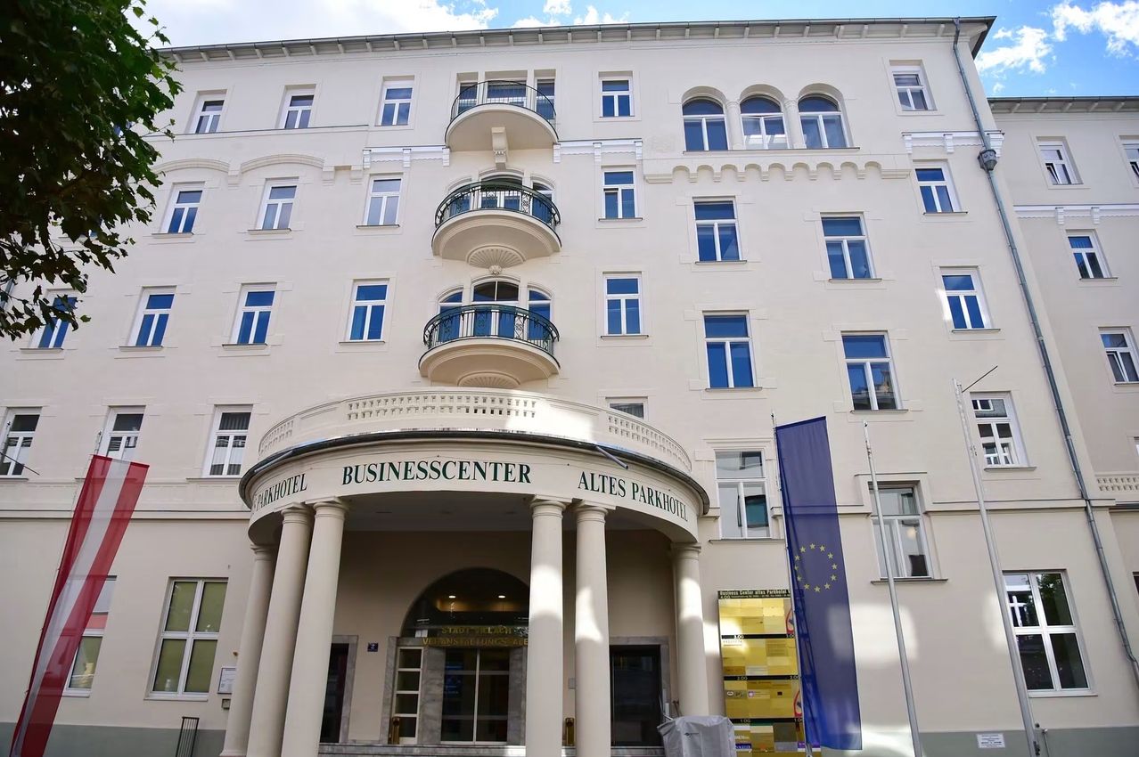 Ein modernes Gebäude mit weißen Wänden, blauen Fenstern und einem Schild, das 'Altes Parkhotel Businesscenter' besagt. Es gibt Säulen und einen Balkon mit Geländern. Eine Flagge befindet sich rechts.