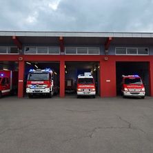 Eine rote Feuerwehrstation mit vier geparkten Feuerwehrwagen im Inneren. Die Wagen sind mit 'Feuerwehr' beschriftet und haben ihre Lichter eingeschaltet. Die Station hat rote Säulen und graue Wände.