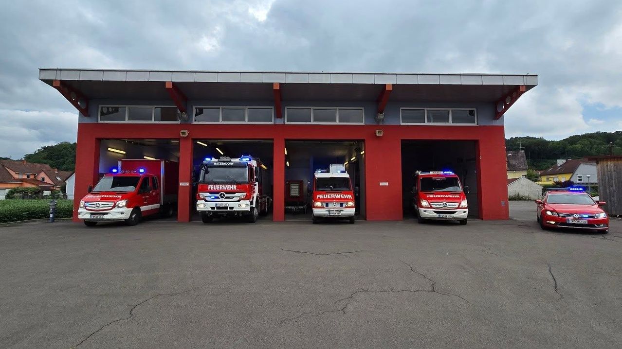 Eine rote Feuerwehrstation mit vier geparkten Feuerwehrwagen im Inneren. Die Wagen sind mit 'Feuerwehr' beschriftet und haben ihre Lichter eingeschaltet. Die Station hat rote Säulen und graue Wände.