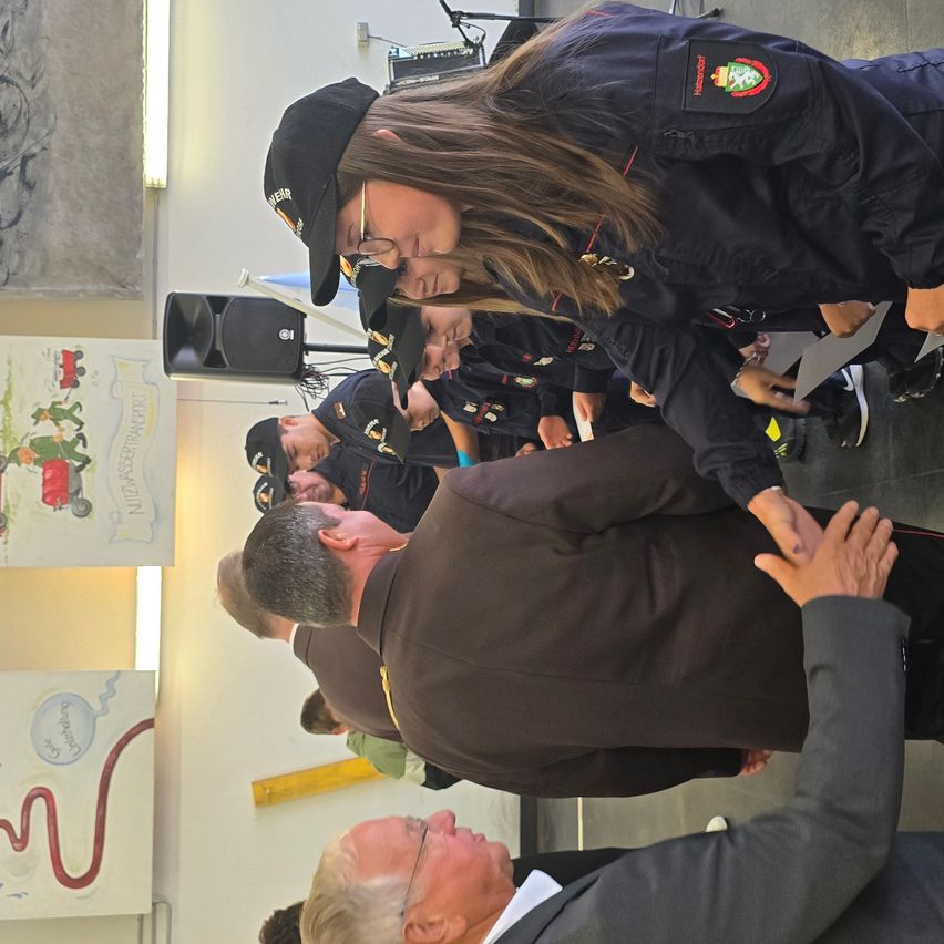 Eine Gruppe von Menschen, möglicherweise Feuerwehrleute, versammelt sich in einem Raum. Eine Frau beugt sich vor und hält die Hand eines Mannes. Sie stehen vor einer Wand mit Zeichnungen.