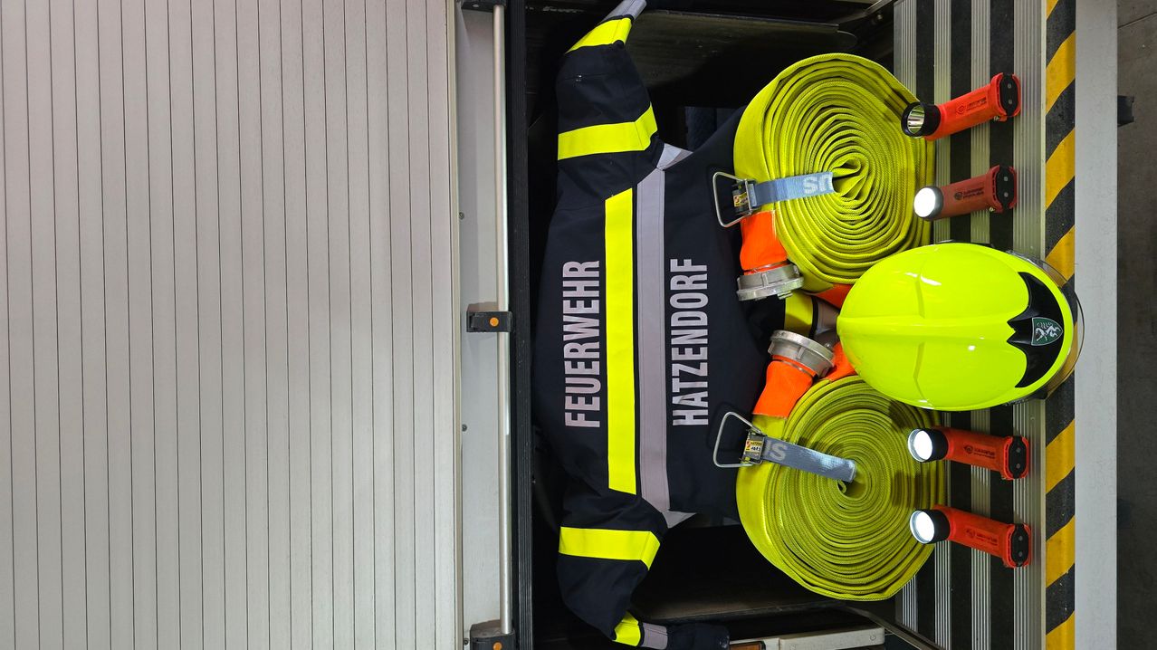 Eine Feuerwehruniform mit reflektierenden Streifen und dem Namen 'Hatzendorf' befindet sich in einem Fahrzeug. Daneben liegen zwei gelbe Feuerwehrschläuche mit 'S' darauf.