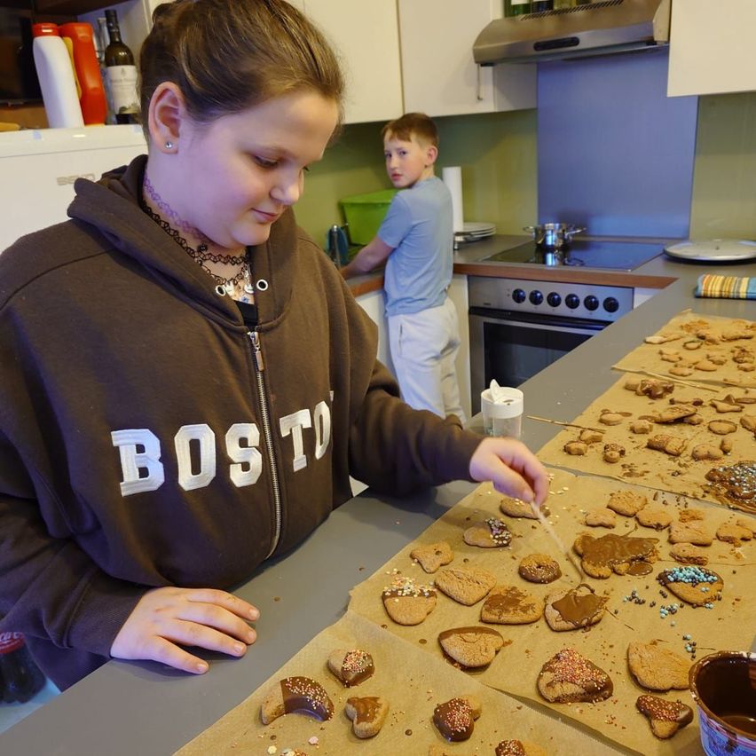 Ein junges Mädchen in einem Boston-Sweatshirt verziert Lebkuchen auf einer Küchentheke, während ein Junge hinter ihr steht.