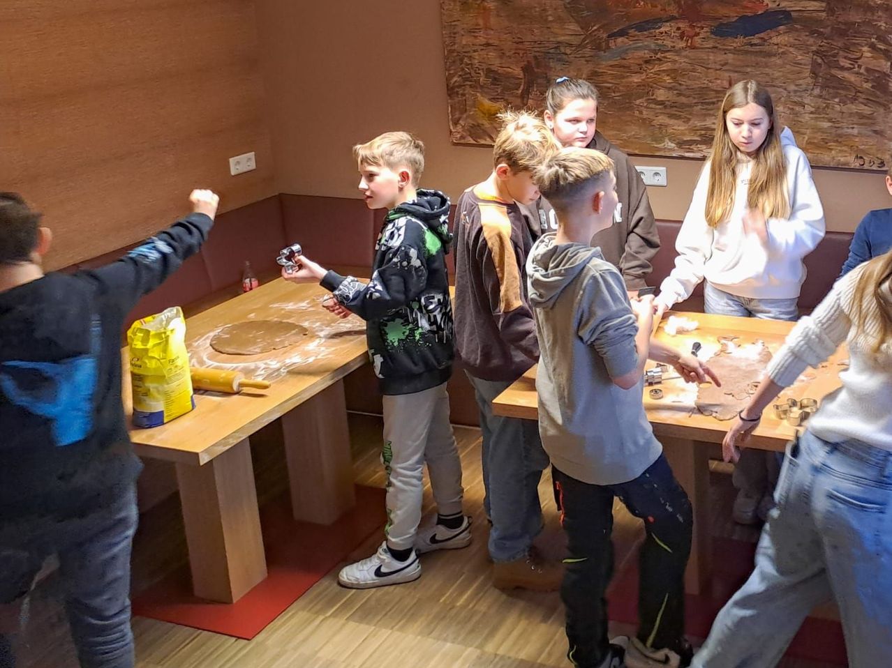 Mehrere Kinder sind um einen Tisch versammelt und machen Teig, während ein anderes Kind auf etwas auf dem Tisch zeigt. Sie befinden sich in einem Raum mit Holzwänden und -böden.