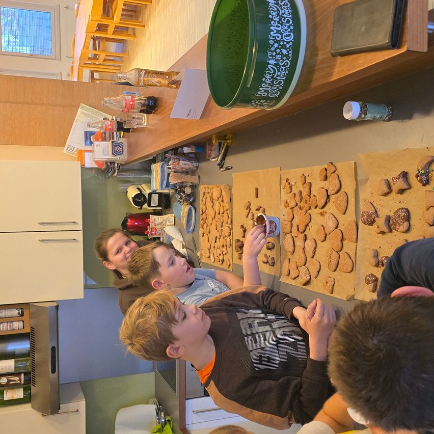 Eine Gruppe von Kindern in einer Küche verziert Kekse, mit einer Frau und einem Jungen hinter ihnen. Verschiedene Kekse liegen auf Papier, und Flaschen und Becher sind auf dem Tresen.