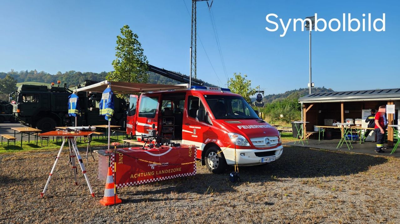 Ein roter Feuerwehrwagen steht in einer Landzone mit einer Vorsichtstafel. Ein Tisch mit einem Drohnenmodell steht in der Nähe.