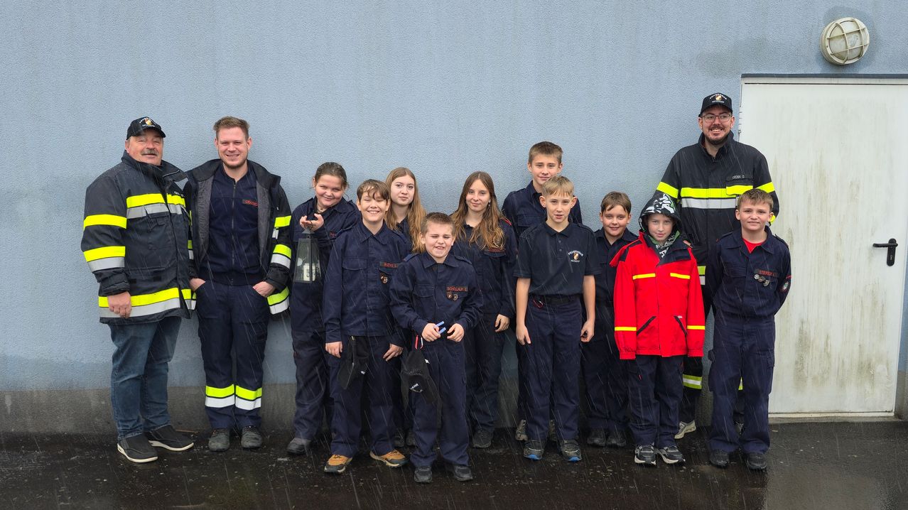 Eine Gruppe junger Menschen in Uniformen und Sicherheitsjacken steht gegen eine blaue Wand und lächelt für ein Foto. Einige halten Helme und eine Laterne.