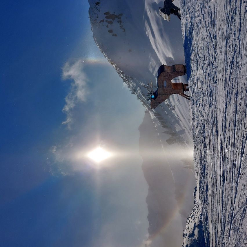 Ein verschneiter Berg mit einem Regenbogen und einer Person in Skiausrüstung am Rande. Die Sonne scheint hell am Himmel.