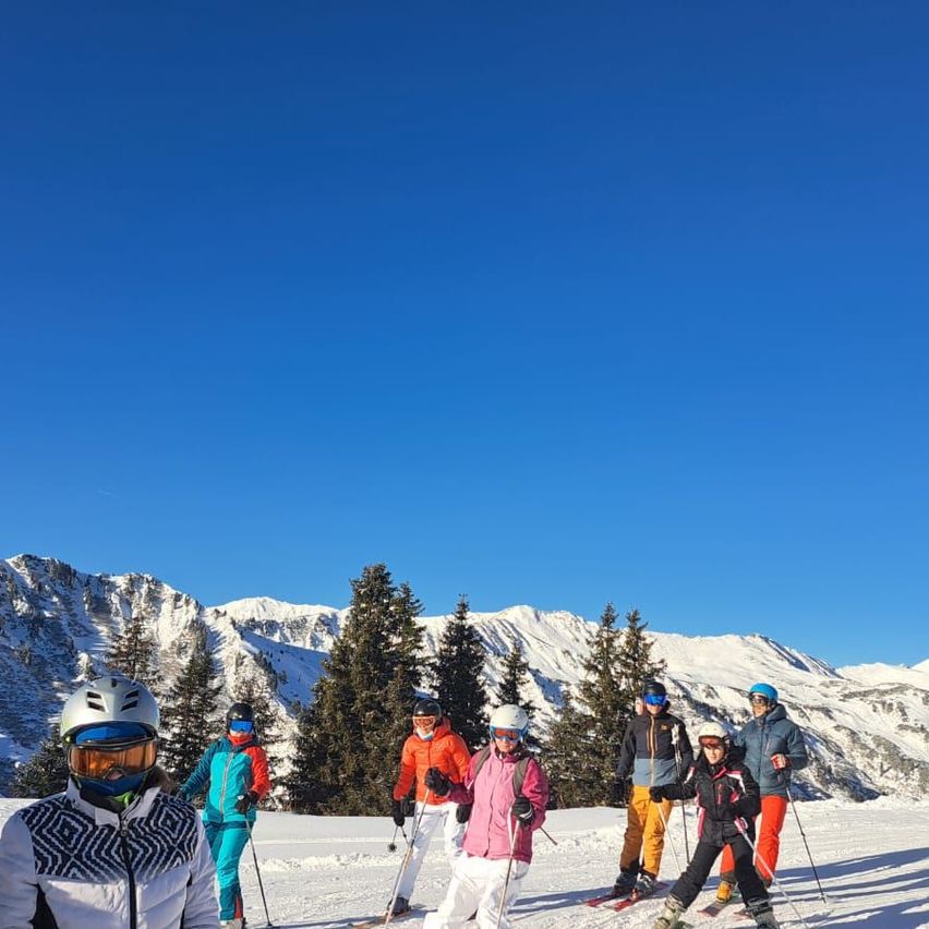 Eine Gruppe von Skifahrern steht auf einem verschneiten Berg, jeder trägt Skiausrüstung und Helme. Sie befinden sich auf einer Piste mit Bäumen und schneebedeckten Bergen im Hintergrund.