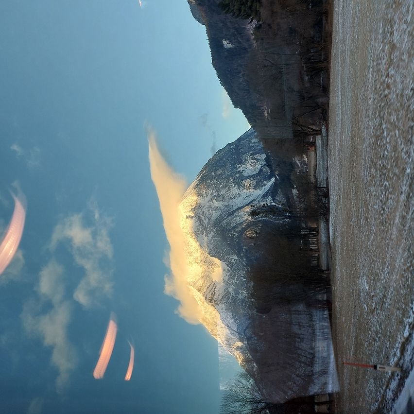Eine Berglandschaft mit schneebedeckten Gipfeln unter einem klaren blauen Himmel. Drei Paraglider sind im Himmel zu sehen.