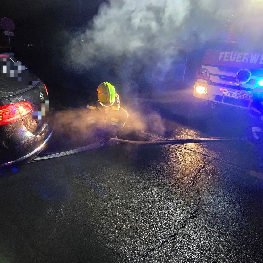 Ein Feuerwehrmann besprüht mit einem Schlauch die Rückseite eines Autos, das Rauch abgibt. Ein Feuerwehrwagen mit blinkenden Lichtern ist in der Nähe geparkt.