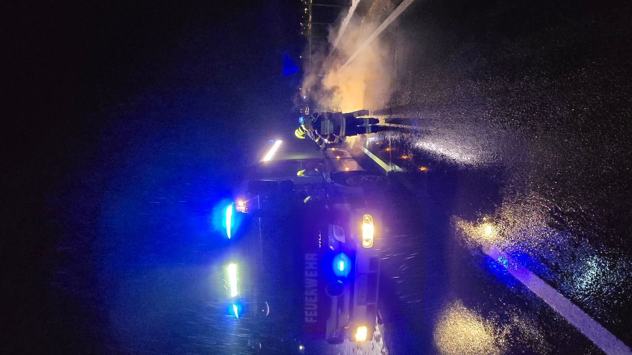 Feuerwehrleute arbeiten bei Nacht auf einer nassen Straße, ein Polizeiauto in der Nähe mit eingeschalteten Lichtern.