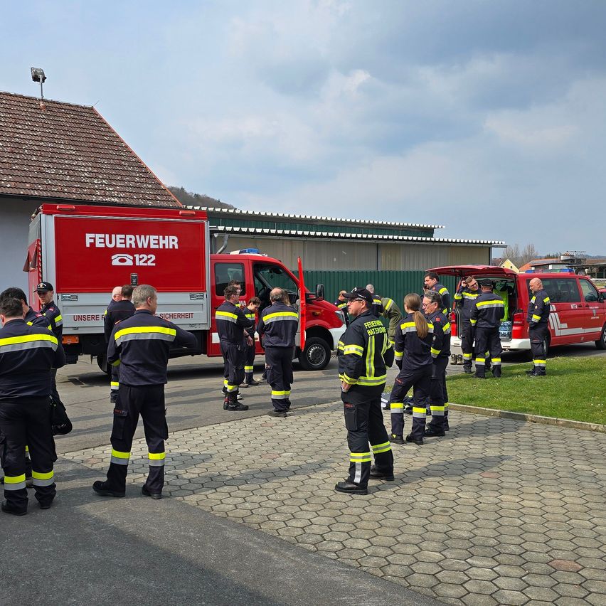 Eine Gruppe Feuerwehrleute steht vor einem roten Feuerwehrwagen mit der Aufschrift 'FEUERWEHR 122'. Sie sind um einen roten Van mit offenem Kofferraum versammelt. Dahinter befindet sich ein Gebäude mit einem braunen Dach.
