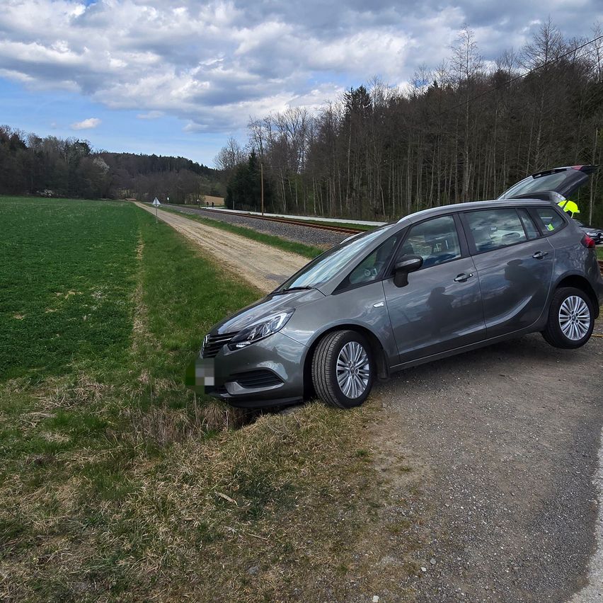 Ein graues Auto mit offenem Kofferraum steht auf einer Feldweg neben einem Grasfeld und einer Eisenbahnstrecke.