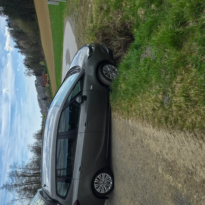 Ein dunkles graues Auto mit geöffneter Hecktür steht auf einem Schotterweg in der Nähe einer Rasenfläche. Das Auto hat ein schlankes Design und befindet sich auf einem leicht gebogenen Weg.