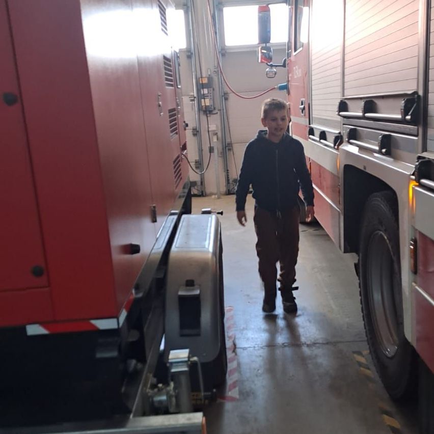 Ein kleiner Junge läuft zwischen zwei roten Feuerwehrautos, eines davon mit einer Leiter an der Seite. Der Bereich ist schwach beleuchtet.