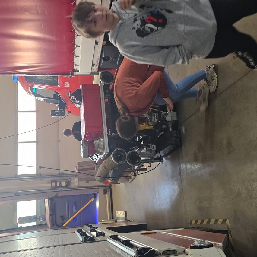 Eine Gruppe von Menschen ist in einer Garage und arbeitet an einem roten Feuerwehrauto. Eine Person ist über den Motor gebeugt, während eine andere in der Nähe steht. Die Garage hat große Fenster und ist mit verschiedenen Geräten gefüllt.