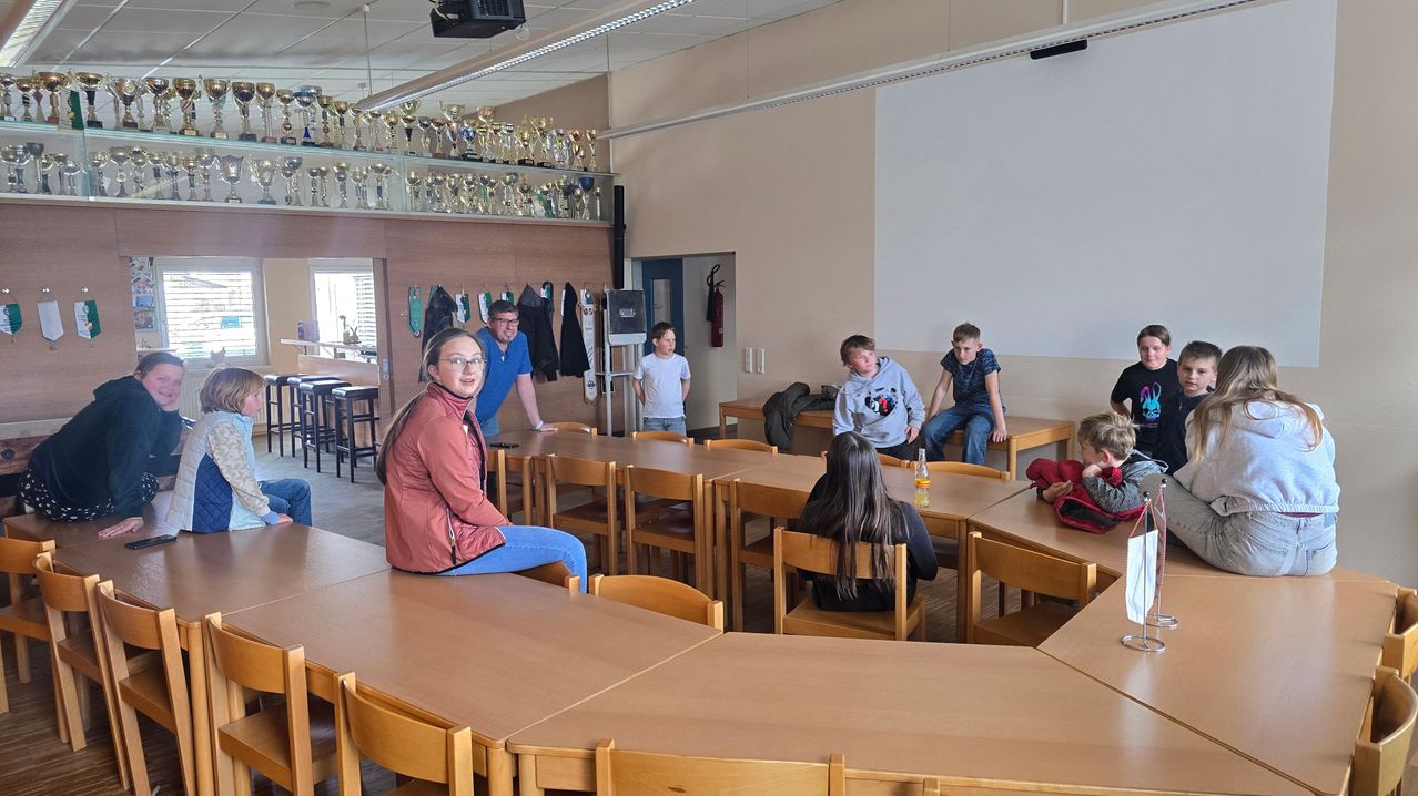 In einem Raum mit Tischen und Stühlen stehen eine Frau und ein Mann in der Nähe eines Tisches. Kinder sitzen auf den Stühlen und Pokale hängen an der Wand. Ein Feuerlöscher ist an der Wand montiert.