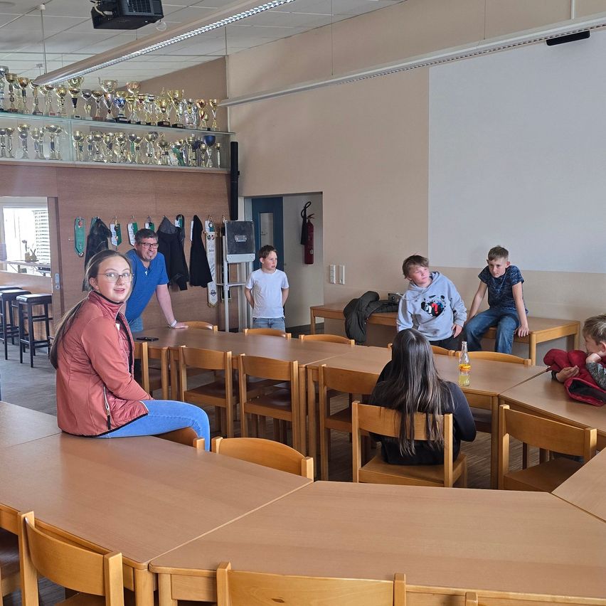 In einem Raum mit Tischen und Stühlen stehen eine Frau und ein Mann in der Nähe eines Tisches. Kinder sitzen auf den Stühlen und Pokale hängen an der Wand. Ein Feuerlöscher ist an der Wand montiert.