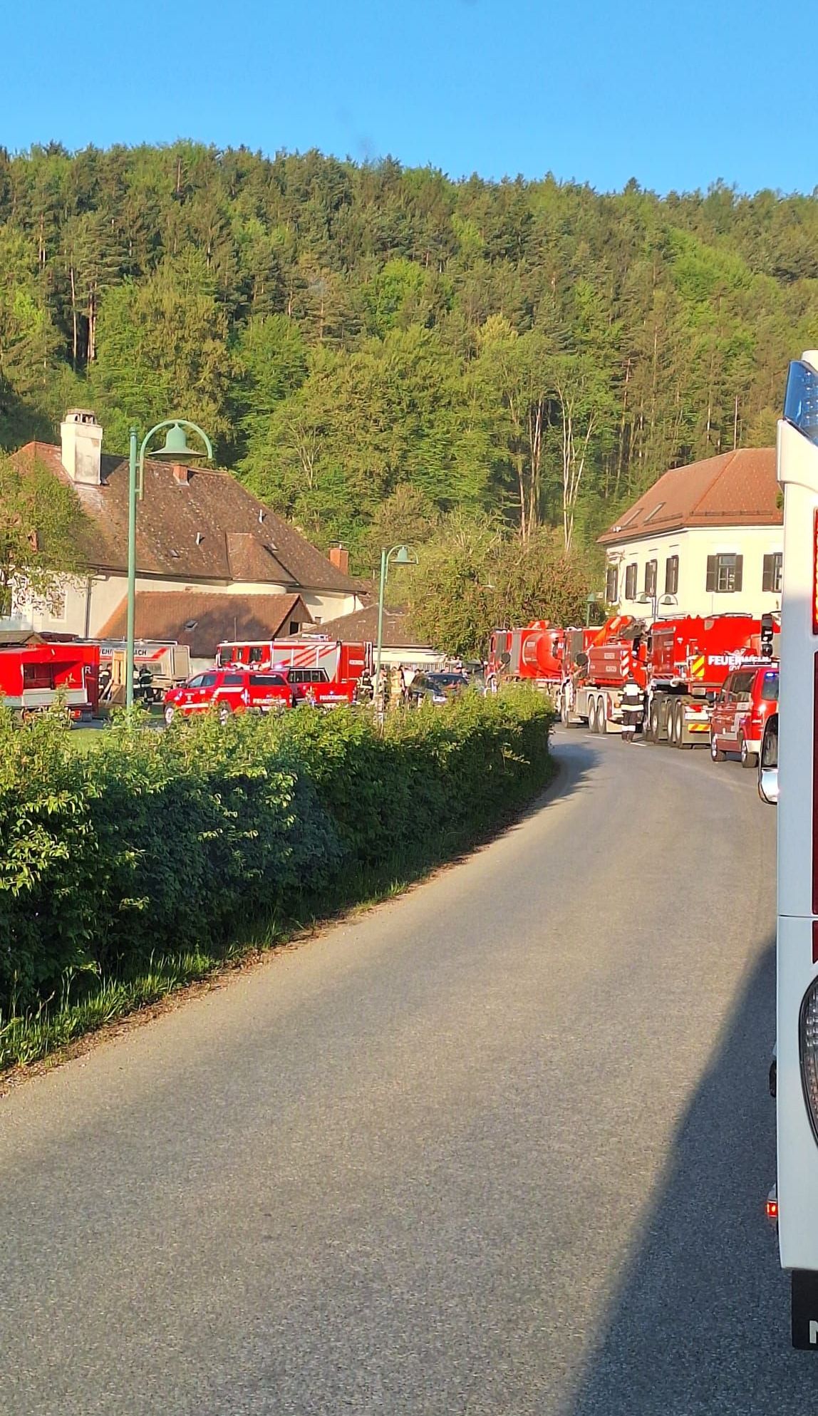 Feuerwehrfahrzeuge sind auf beiden Seiten einer Straße geparkt, mit mehreren Personen um sie herum. Ein Gebäude mit Schornstein ist auf der linken Seite.