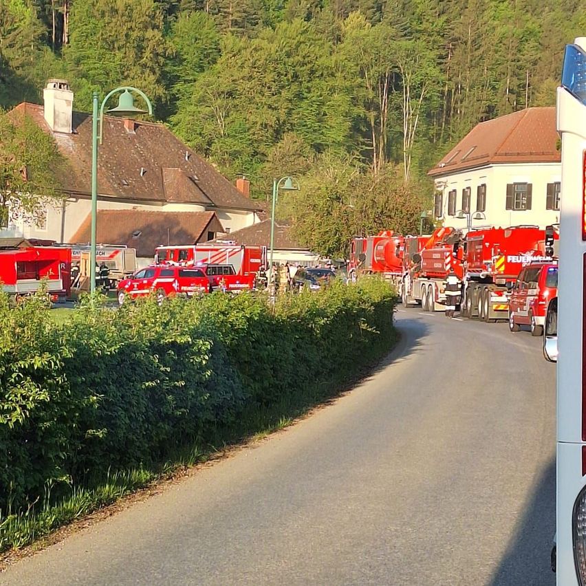 Feuerwehrfahrzeuge sind auf beiden Seiten einer Straße geparkt, mit mehreren Personen um sie herum. Ein Gebäude mit Schornstein ist auf der linken Seite.