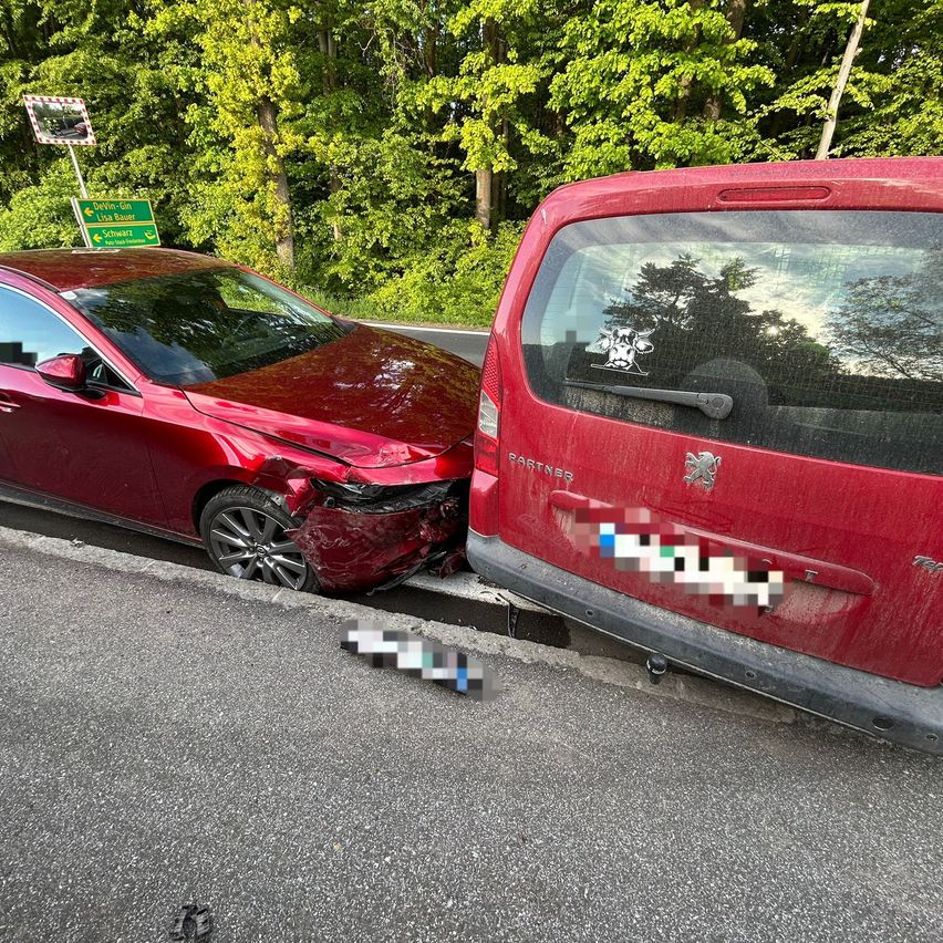 Zwei rote Autos sind in einen Unfall verwickelt, wobei der Stoßfänger eines Autos beschädigt ist und das andere am Straßenrand geparkt ist.