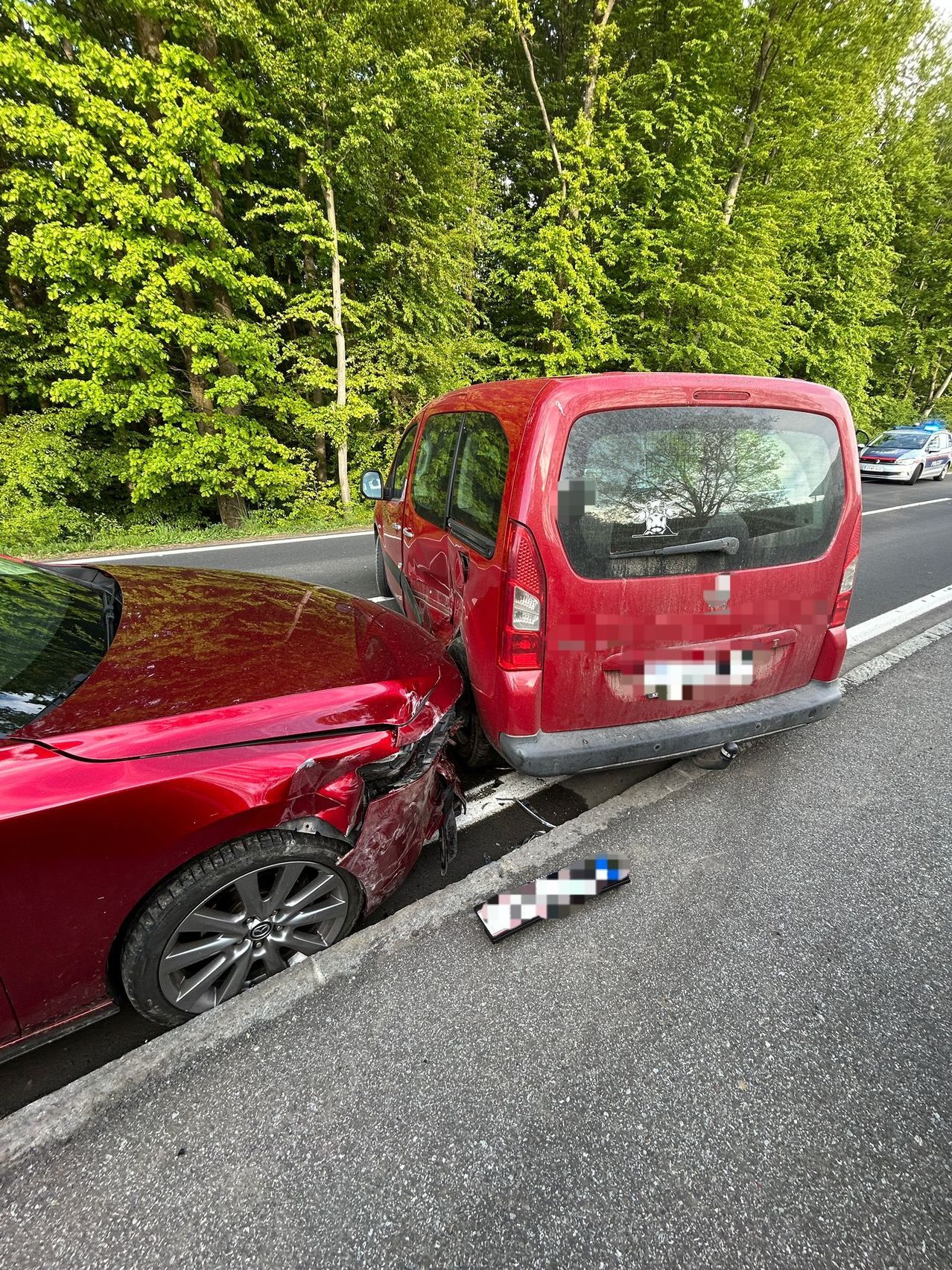 Zwei rote Autos sind am Straßenrand geparkt, eines mit einem beschädigten Stoßfänger. Im Hintergrund sind Bäume und ein Polizeiauto zu sehen.