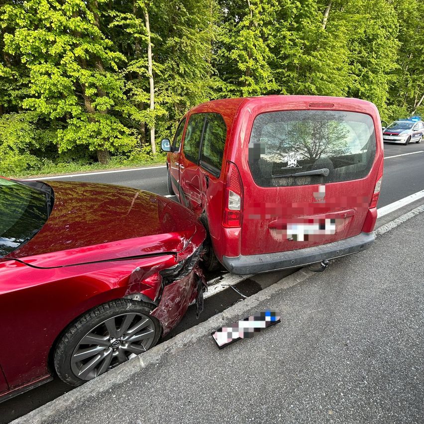Zwei rote Autos sind am Straßenrand geparkt, eines mit einem beschädigten Stoßfänger. Im Hintergrund sind Bäume und ein Polizeiauto zu sehen.