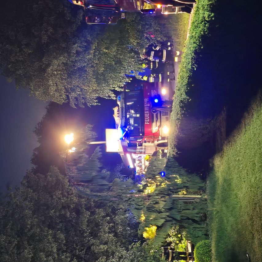 Feuerwehrleute in voller Montur kümmern sich um einen Notfall in der Nähe eines Feuerwehrwagens mit blinkenden Lichtern und einem rot-weißen Farbschema. Bäume und ein Zaun umgeben die Szene bei Nacht.