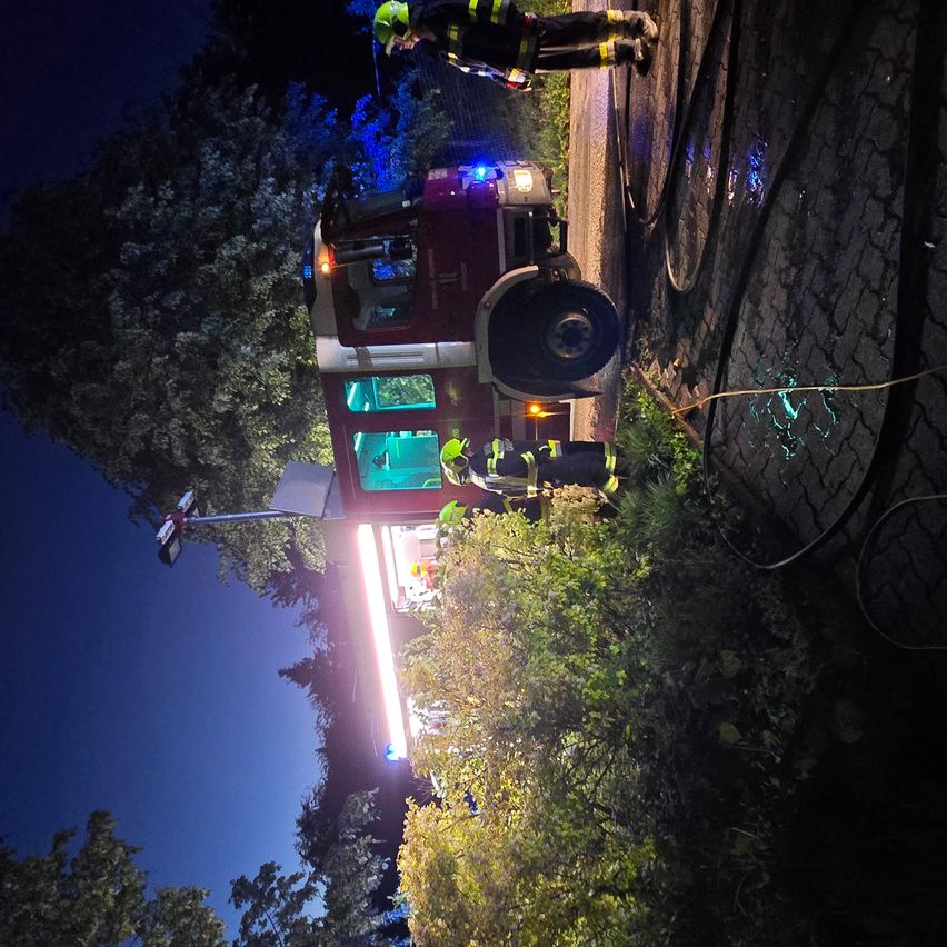 Feuerwehrleute in voller Montur stehen neben einem nachts geparkten Feuerwehrauto. Sie halten einen mit dem Auto verbundenen Schlauch, mit Lichtern und Bäumen im Hintergrund.