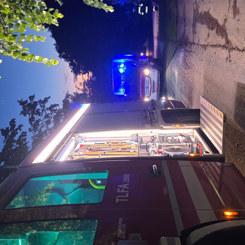 Eine obere Ansicht eines Feuerwehrwagens mit offener Hecktür, blinkenden Lichtern, geparkt am Straßenrand in der Dämmerung.