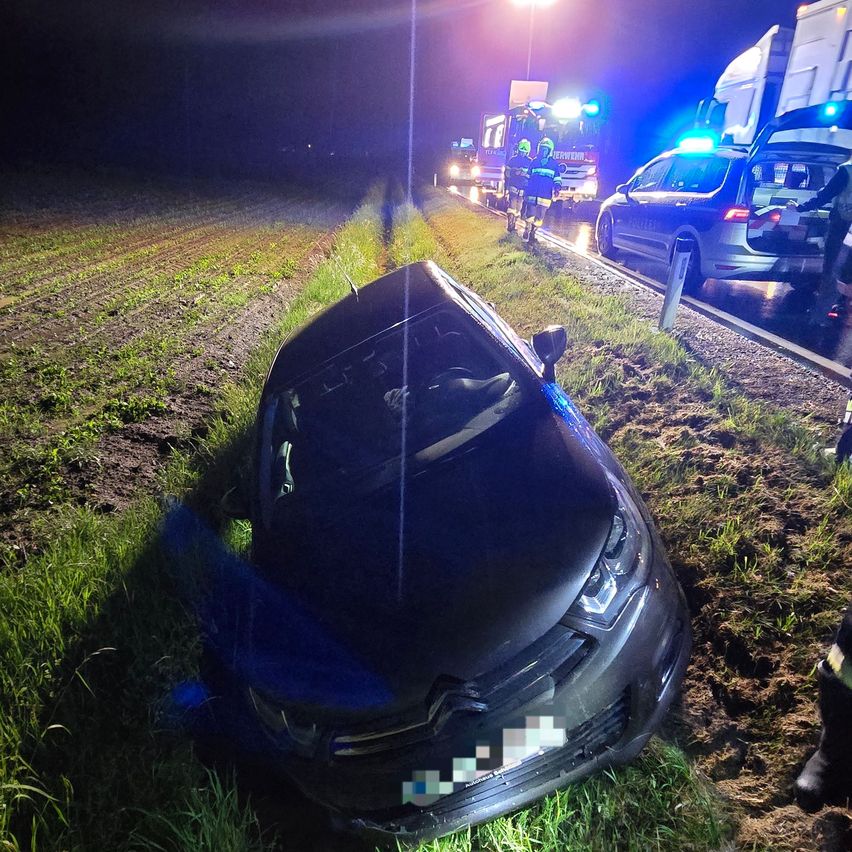 Ein blaues Auto ist nachts auf der Seite einer Straße zerstört. Rettungsdienste sind anwesend und ihre Lichter sind eingeschaltet.