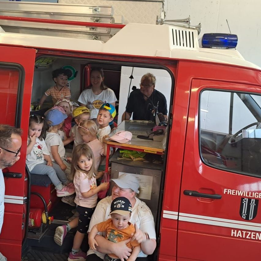 Eine Gruppe von Kindern und Erwachsenen befindet sich in einem roten Feuerwehrauto mit offener Tür. Ein Mann hält ein Baby und sitzt mit einem anderen Mann. Ein Mann spricht in ein Mikrofon im Inneren des Fahrzeugs.