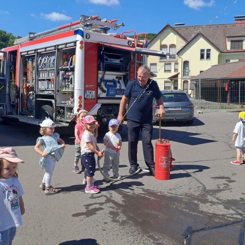 Ein älterer Mann hält einen Feuerlöscher in der Hand und steht vor einem Feuerwehrauto. In der Nähe stehen Kinder und beobachten den Mann. Im Hintergrund befinden sich Häuser und ein Auto.