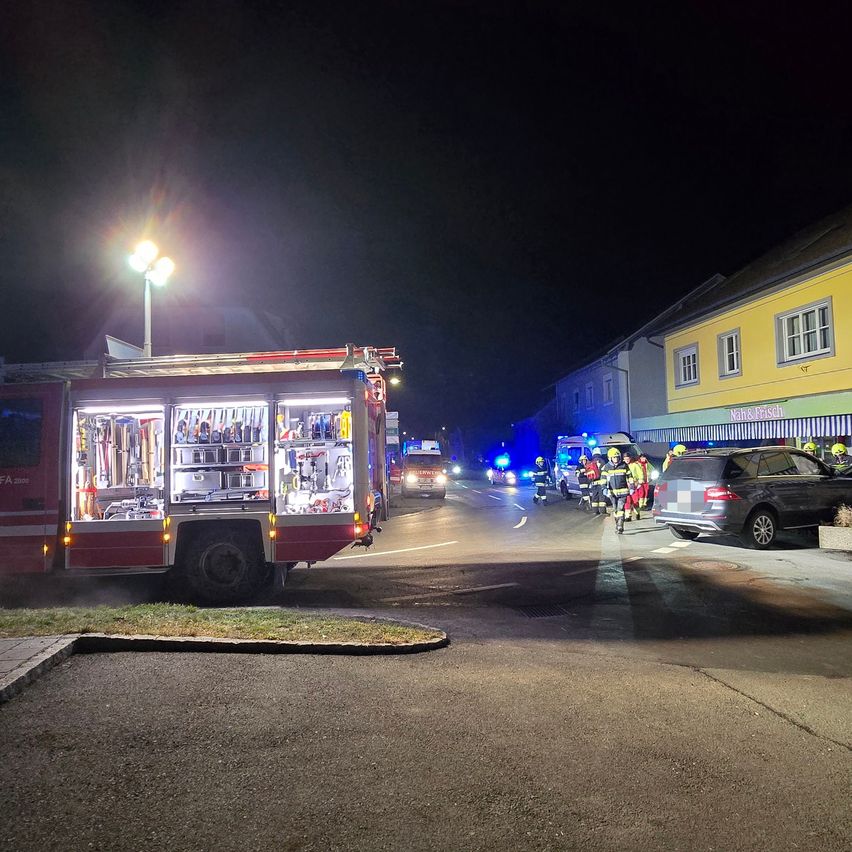 Rettungsfahrzeuge, einschließlich eines Feuerwehrwagens, sind auf der Straße geparkt. Mehrere Feuerwehrleute kümmern sich um einen Vorfall, wobei blinkende blaue Lichter die Szene beleuchten.