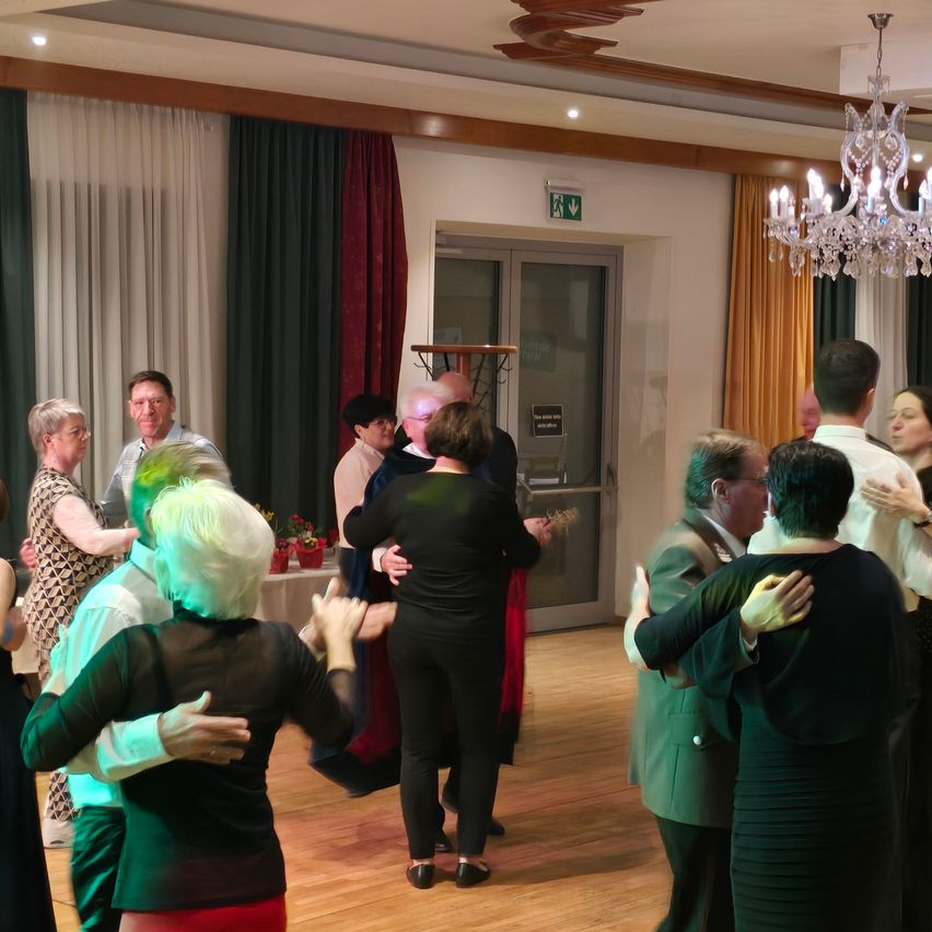 Eine Gruppe von Menschen tanzt in einem Saal. Ein Kronleuchter und Vorhänge schmücken den Raum. Einige tragen Brillen und formelle Kleidung. Eine Gitarre ruht auf einem Ständer.