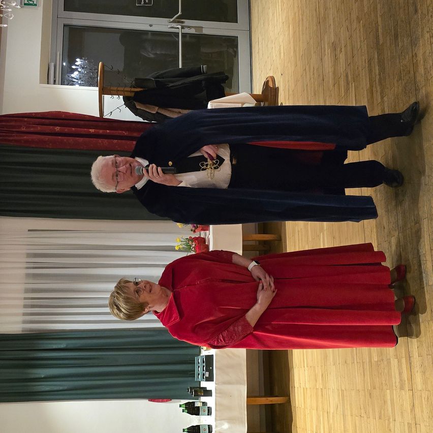 Ein Mann und eine Frau stehen auf einem Holzboden und sehen sich an. Der Mann hält ein Mikrofon und scheint zu sprechen. Die Frau trägt ein rotes Kleid und eine Brille. Hinter ihnen befindet sich ein weißer Vorhang und ein Tisch mit Flaschen.