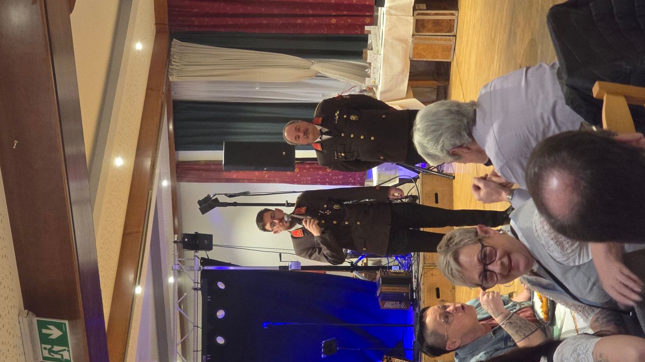 Ein Soldat in Uniform spricht in ein Mikrofon. Ein anderer Offizier steht hinter ihm. Zwei Personen, eine mit Brille, lauschen aufmerksam. Die Szene umfasst eine Bühne mit blauem Hintergrund, Lautsprechern und einem Holzboden.