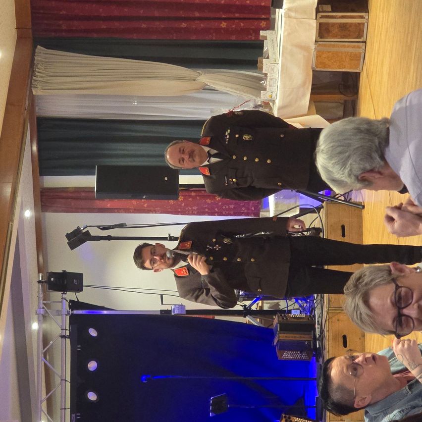 Ein Soldat in Uniform spricht in ein Mikrofon. Ein anderer Offizier steht hinter ihm. Zwei Personen, eine mit Brille, lauschen aufmerksam. Die Szene umfasst eine Bühne mit blauem Hintergrund, Lautsprechern und einem Holzboden.