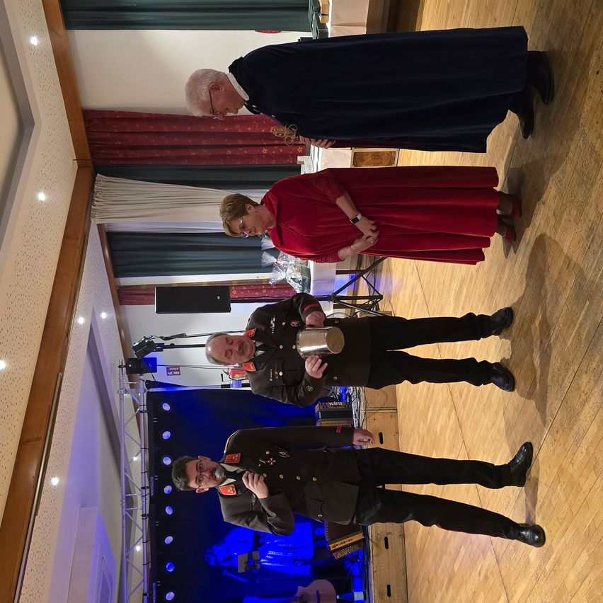 Mehrere Menschen sind drinnen versammelt, möglicherweise für eine formelle Veranstaltung. Ein älterer Mann und eine Frau in einem roten Kleid stehen zusammen, während zwei Männer in Militäruniformen unter ihnen stehen. Einer der Männer hält einen silbernen Becher.