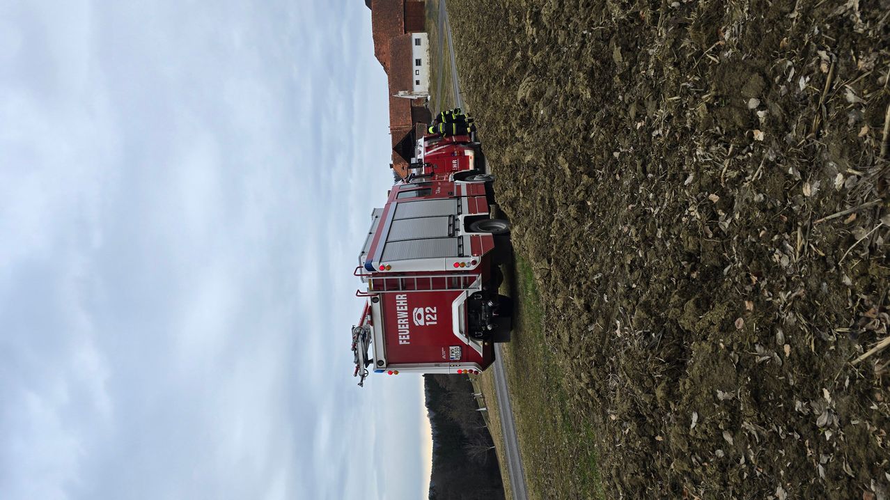 Ein roter Feuerwehrwagen mit der Nummer 122 steht am Straßenrand. Zwei Feuerwehrleute in gelber Schutzkleidung stehen neben dem Wagen.