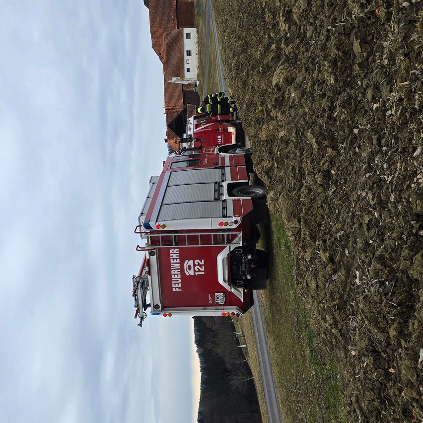 Ein roter Feuerwehrwagen mit der Nummer 122 steht am Straßenrand. Zwei Feuerwehrleute in gelber Schutzkleidung stehen neben dem Wagen.
