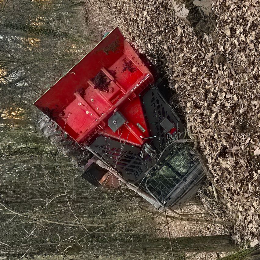 Ein roter Maschinenwagen, wahrscheinlich ein Radlader, ist in einem Wald umgekippt, wobei Blätter das Gebiet bedecken.