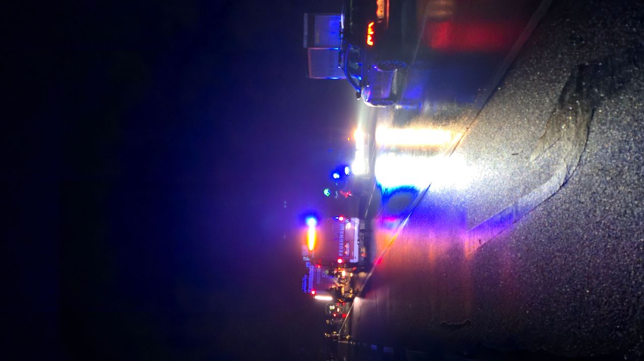 Einsatzfahrzeuge stehen mit blinkenden Lichtern am Straßenrand. Der Bereich ist dunkel und die Straßenoberfläche ist nass.