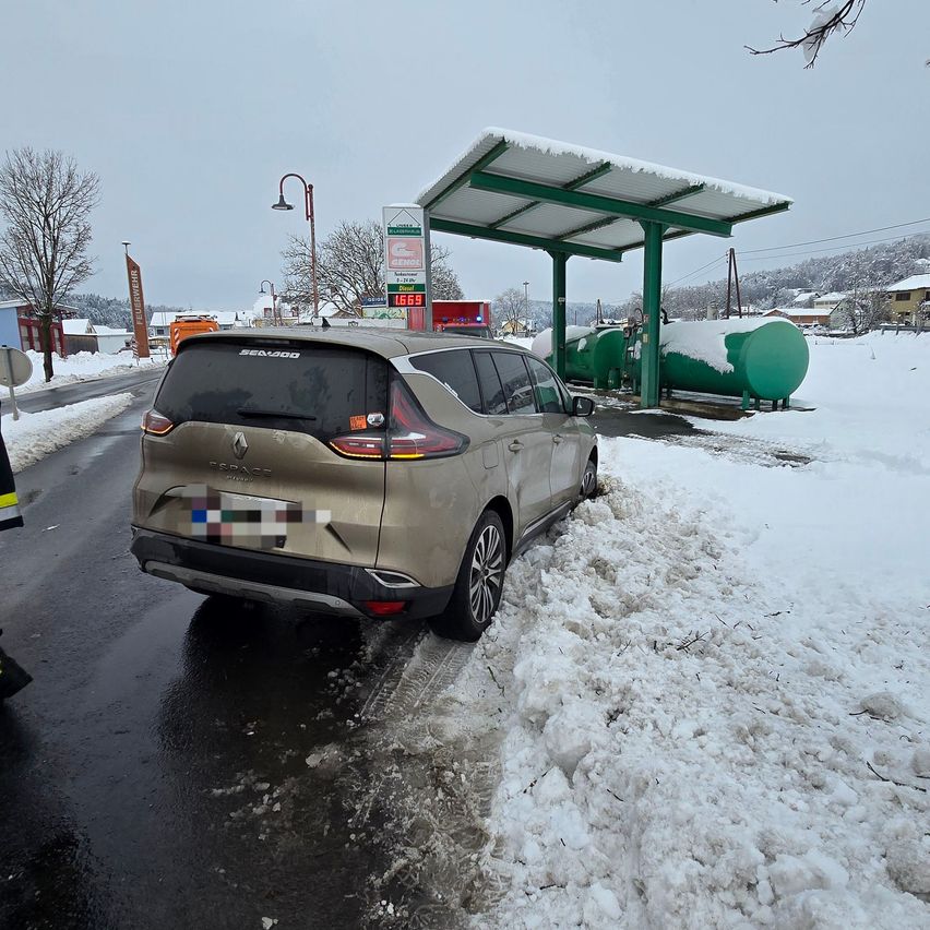 Ein beigefarbener Renault SUV ist auf einer verschneiten Straße geparkt, in der Nähe einer Tankstelle mit einem grünen Tank und einem Vordach. Schnee bedeckt den Boden und einen kahlen Baum.