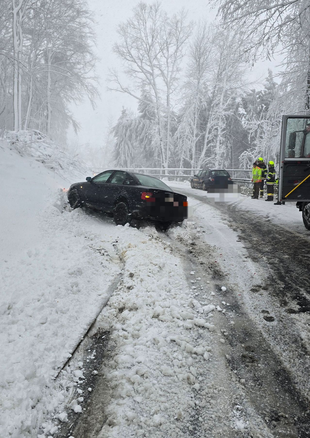 Ein schwarzes Auto steckt im Schnee auf einer verschneiten Straße fest. Zwei andere Autos sind in der Nähe geparkt. Zwei Personen in reflektierenden Westen stehen bei einem Fahrzeug.