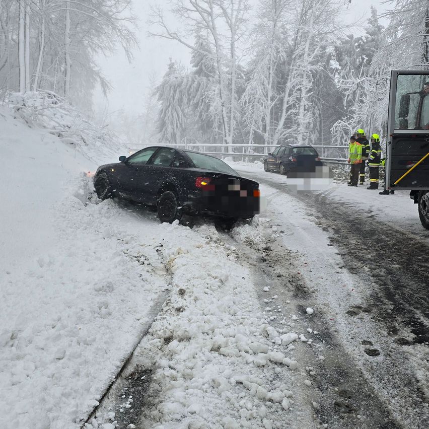 Ein schwarzes Auto steckt im Schnee auf einer verschneiten Straße fest. Zwei andere Autos sind in der Nähe geparkt. Zwei Personen in reflektierenden Westen stehen bei einem Fahrzeug.