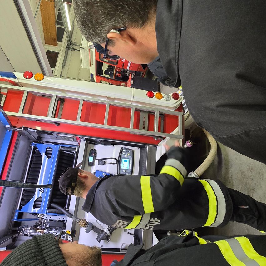 Zwei Feuerwehrleute arbeiten an einem Feuerwehrauto, einer hält einen Schlauch und der andere arbeitet an der Ausrüstung. Eine Leiter ist am Auto montiert.