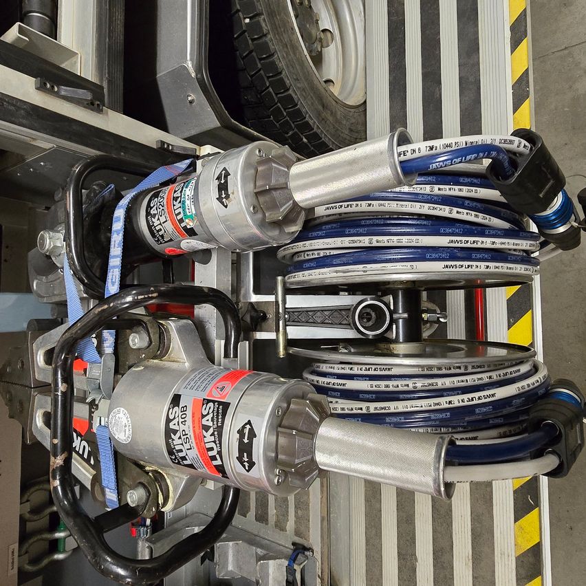 Eine Nahaufnahme von zwei hydraulischen Jacken, die an einem Metallrahmen befestigt sind, mit blauen Schläuchen, die darum gewickelt sind. Ein Reifen ist im Hintergrund zu sehen.