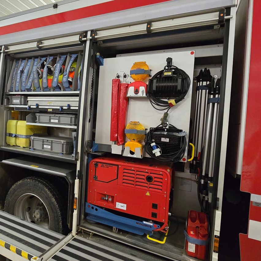 Der Innenraum eines Feuerwehrfahrzeuges ist offen und zeigt einen roten Generator und eine Sammlung von Werkzeugen und Ausrüstung. Aufbewahrungsboxen und Container sind auf den Regalen.